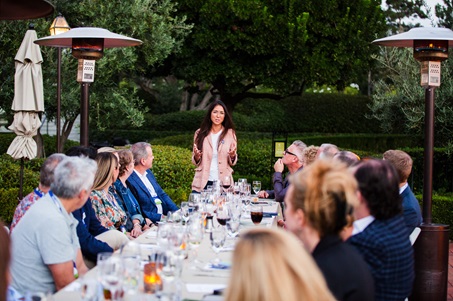 La cuidadora Caresse Sakagawa de pie en la cabecera de la mesa hablando con una larga mesa de invitados.